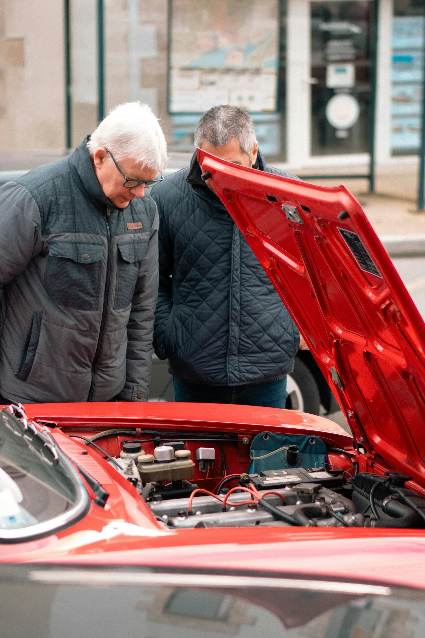Deux hommes penchés au-dessus du capot ouvert d'une voiture rouge garée dans une rue, en train d'examiner le moteur et d'en discuter.